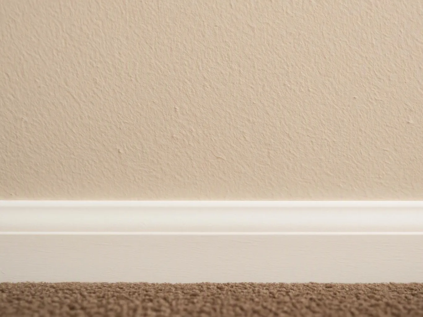 A beige wall with white baseboard meets brown carpet in a simple indoor corner