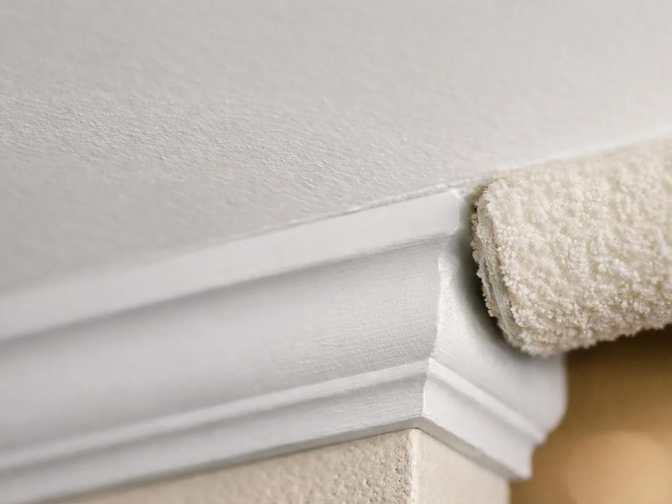 A paint roller applies white paint to the top edge of a wall and trim corner