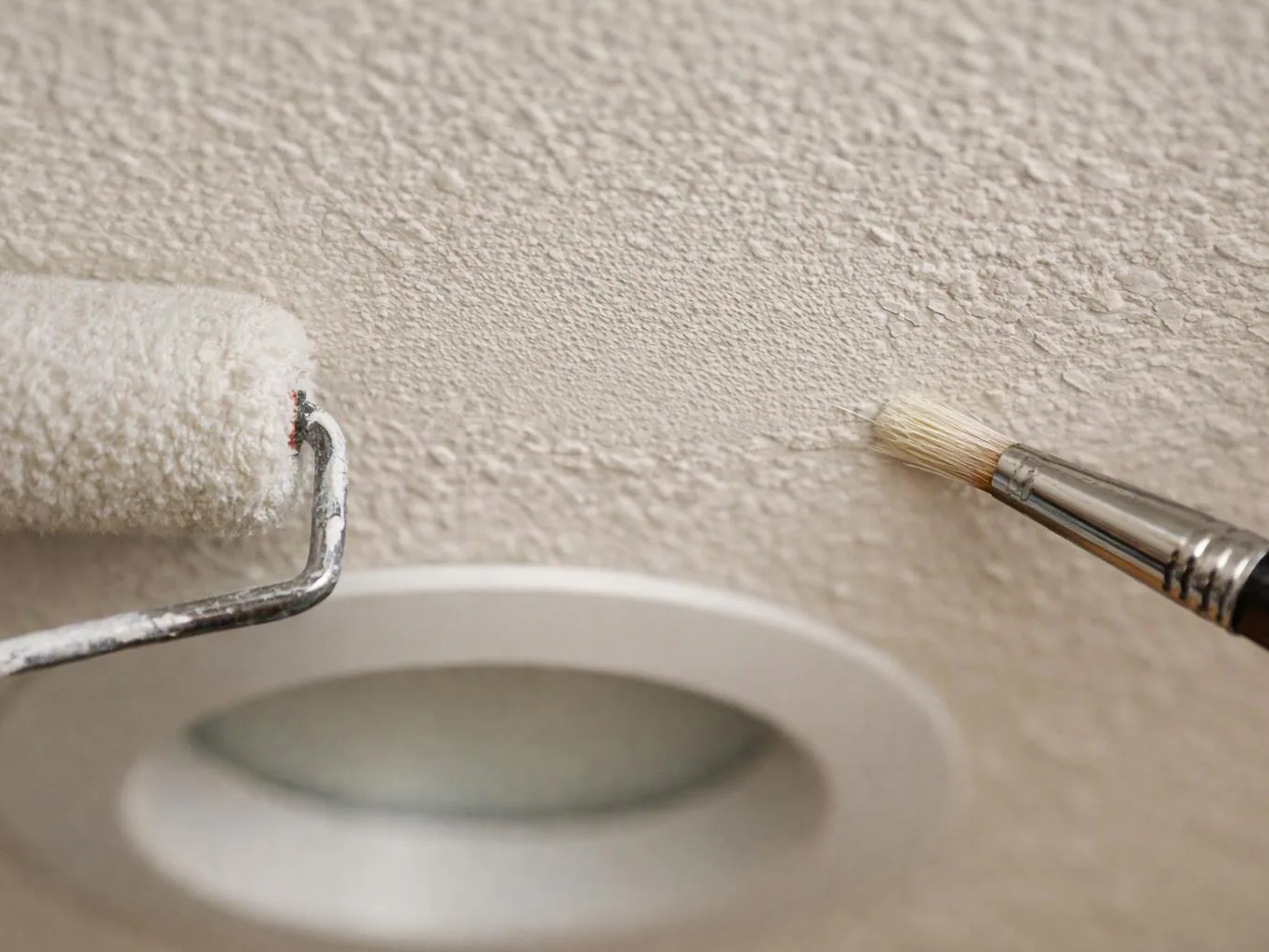 A paint roller and small brush apply white paint around a recessed ceiling light opening