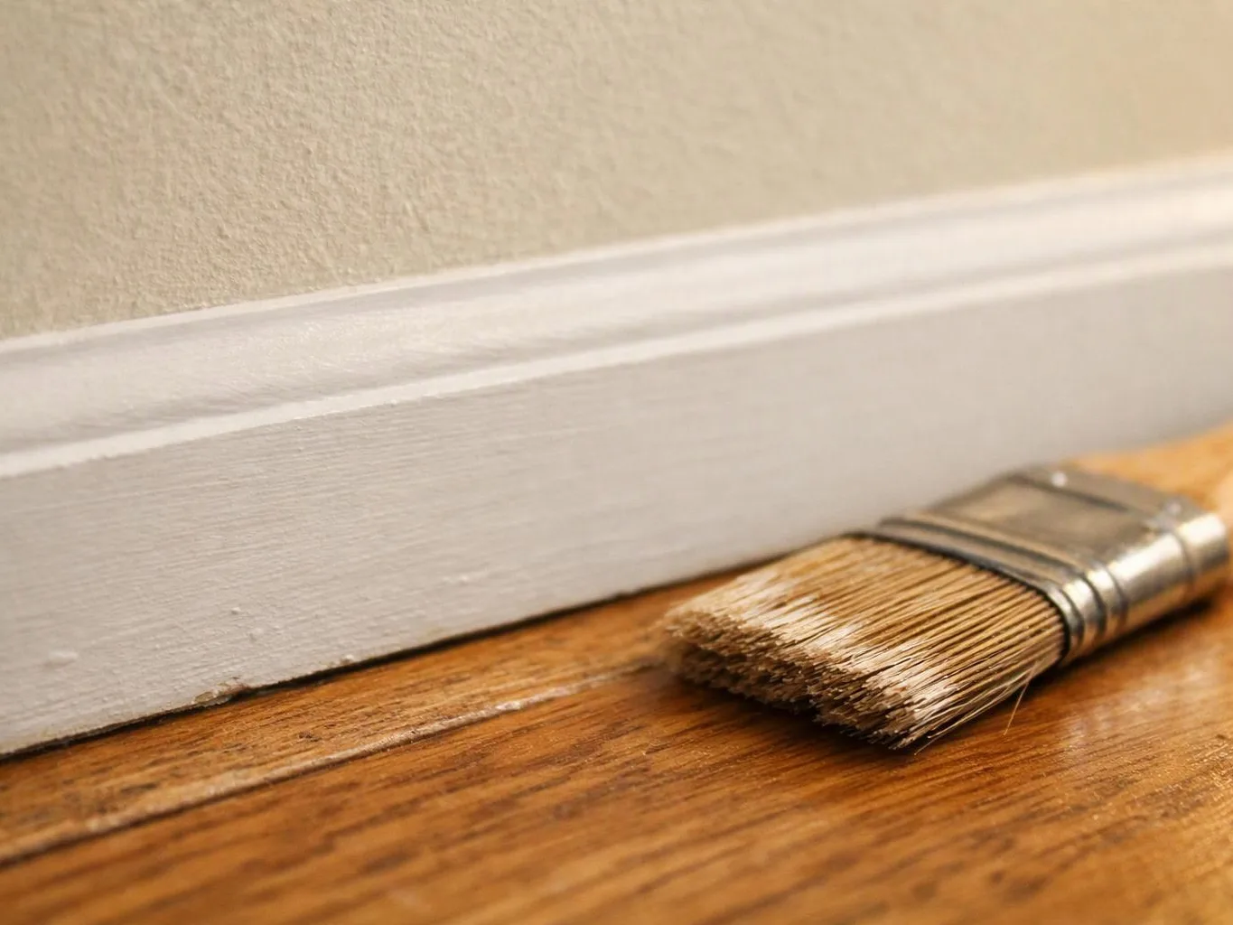 A paintbrush rests on a wooden floor beside a freshly painted white baseboard and wall